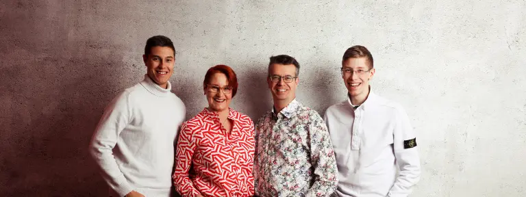 3 Männer und eine Frau stehen nebeneinander vor einer grauen Wand und lächeln