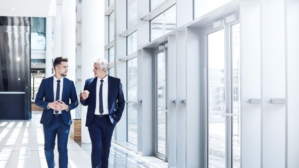 2 Männer in blauem Anzug und Krawatte laufen durch ein Bürogebäude und unterhalten sich