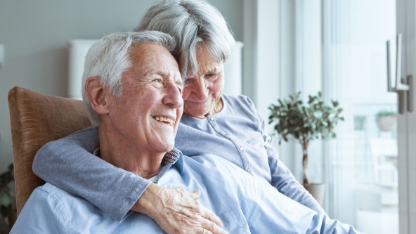 Ältere Frau hat älteren Mann neben sich im Arm - beide sitzen auf einem Sessel und lachen zusammen