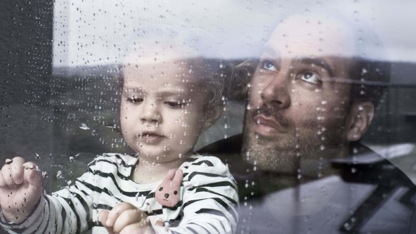 Junger Mann steht hinter einem Kleinkind - gemeinsam schauen sie aus einem Fenster, auf dem Regentropfen zu sehen sind