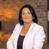 Frau mit dunklen Haaren und rosa Kostüm sitzt auf einem Stuhl