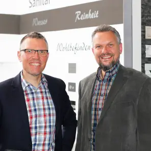 Zwei Männer, einer davon mit Brille, stehen nebeneinander und lächeln