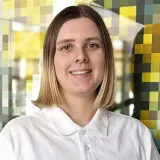 Junge Frau mit kurzen blonden Haaren und weißem Poloshirt steht vor bunter Wand