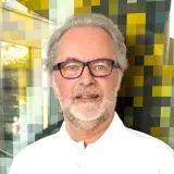 Mann mit kurzen grauen Haaren, Brille und weißem Poloshirt steht vor bunter Wand