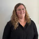 Frau mit blonden lockigen Haaren und Brille steht vor einer Wand und lächelt