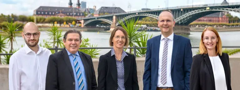 Drei Männer und zwei Frauen in Business-Kleidung vor einem Fluss mit Brücke