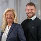 Frau mit blonden Haaren und Blazer und junger Mann mit Pullover stehen lächelnd nebeneinander