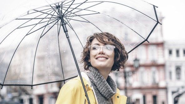 Frau mit Brille und gelber Jacke hat einen durchsichtigen Regenschirm aufgespannt