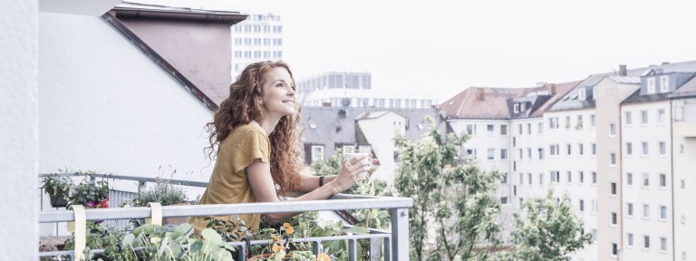 Frau steht auf Balkon mit Kaffeetasse in der Hand und blickt in die Ferne