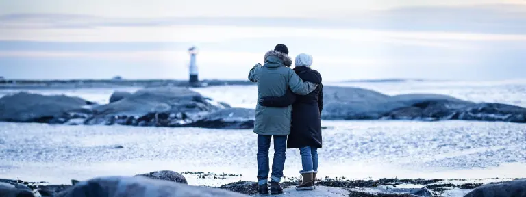 Mann und Frau stehen mit Jacken und Mützen am Strand und blicken auf einen Leuchtturm