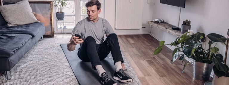 Junger Mann sitzt auf Sportmatte und hat Smartphone in der Hand