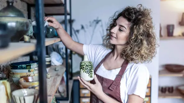 Junge Frau mit lockigen Haaren und einer roten Schürze räumt Tongegenstände in ein Regal
