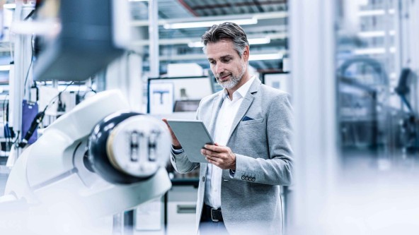 Mann mit Bart und in hellem Anzug schaut in einer Maschinenhalle auf sein Tablet