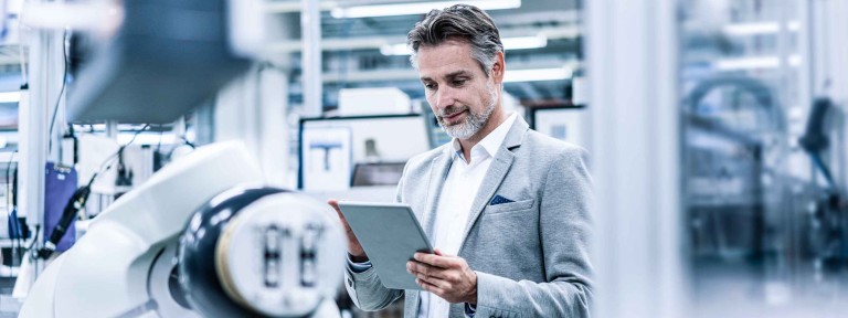 Mann mit Bart und in hellem Anzug schaut in einer Maschinenhalle auf sein Tablet