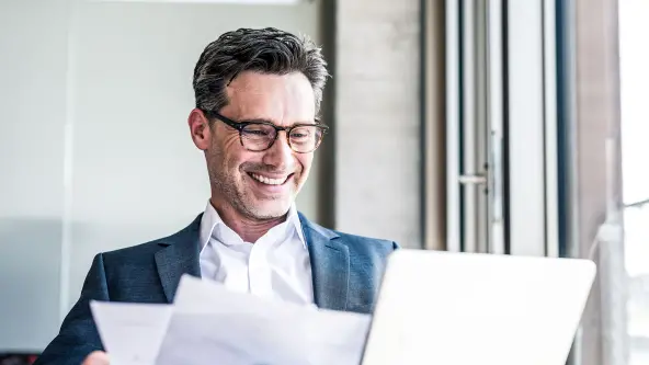 Mann mit Brille blickt lachend auf Tablet und Unterlagen