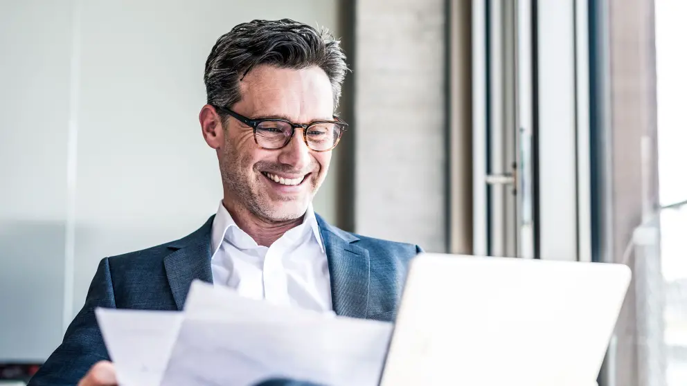 Mann mit Brille blickt lachend auf Tablet und Unterlagen
