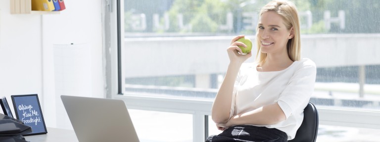Junge Frau hat grünen Apfel in der Hand und sitzt vor einem aufgeklappten Laptop