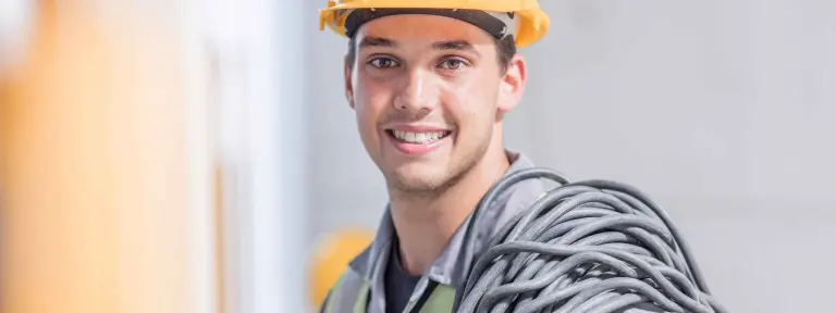 Junger Elektroniker mit Helm und Kabeln lächelt direkt in die Kamera