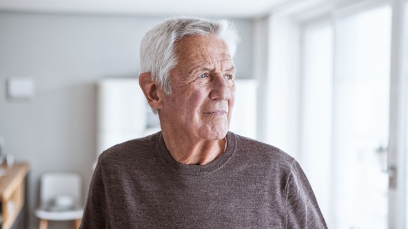 Älterer Mann blickt skeptisch aus Fenster seiner Wohnung
