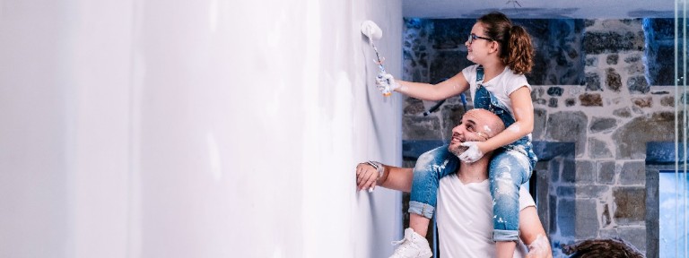 Ein Mann trägt ein Mädchen auf seinen Schultern und sie streicht die Wand mit weißer Farbe