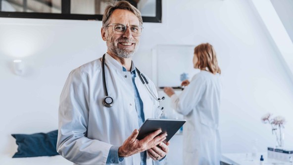 Ein Arzt mit Brille hält Tablet in der Hand und im Hintergrund steht eine Helferin