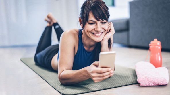 Junge Frau liegt auf Trainingsmatte und lacht in ihr Smartphone