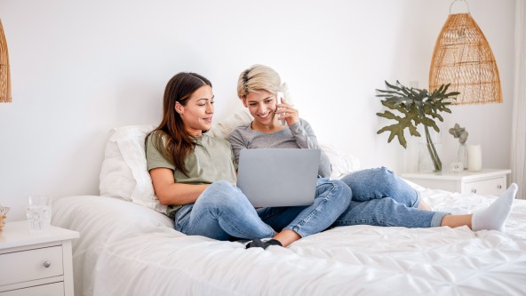 2 junge Frauen sitzen auf einem Bett mit Telefon und Laptop