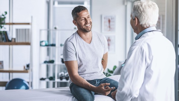 Lächelnder Mann sitzt auf einer Liege, vor ihm steht ein Mann im Arztkittel