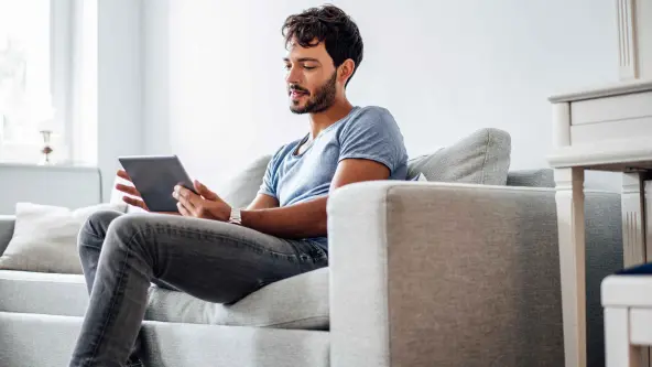 Junger Mann sitzt mit einem Tablet in der Hand auf einem grauen Sofa
