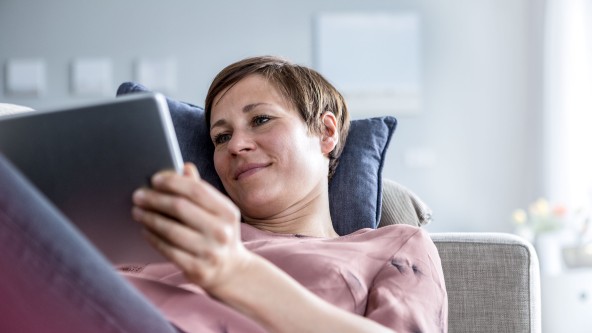 Frau liegt auf Couch und schaut grinsend in ihr Tablet