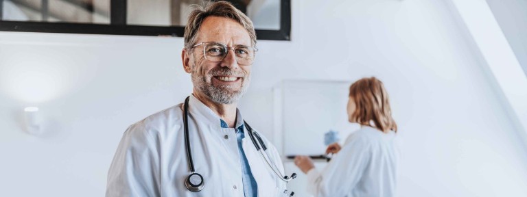 Ein Arzt mit Brille hält Tablet in der Hand und im Hintergrund steht eine Helferin