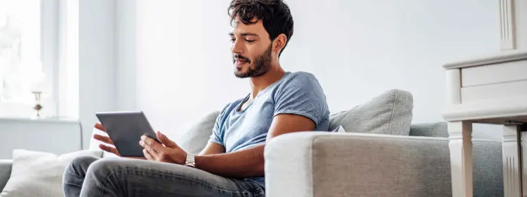 Junger Mann sitzt mit einem Tablet in der Hand auf einem grauen Sofa