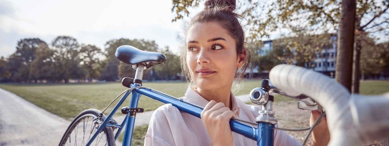 Frau trägt blaues Fahrrad auf Schulter durch einen Park