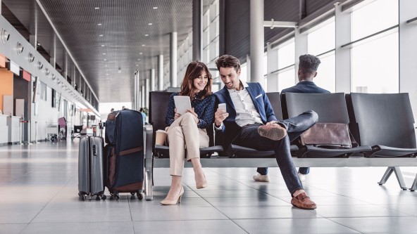 Mann mit Handy in der Hand sitzt neben einer Frau mit Tablet in der Hand im Flughafen