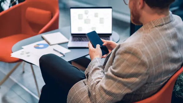 Rückenansicht eines Mannes, der sein Smartphone in der Hand hält, im Hintergrund auf einem Tisch steht sein offener Laptop