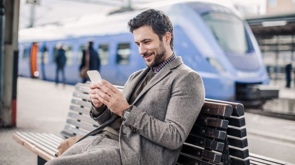 Mann sitzt mit Smartphone in der Hand auf Bank am Bahnhof