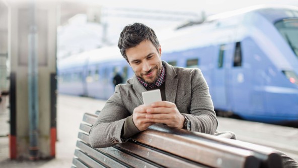 Mann sitzt mit Smartphone in der Hand auf einer Bank am Bahnhof