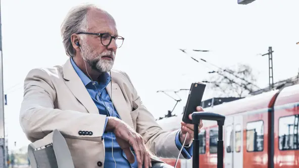 Älterer Mann mit Brille blickt am Bahnhof in sein Handy