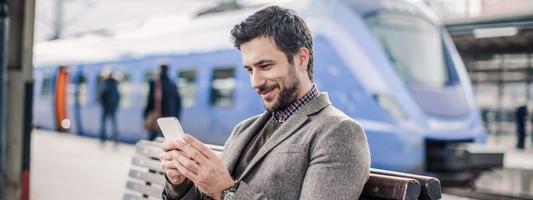 Mann sitzt mit Smartphone in der Hand auf Bank am Bahnhof