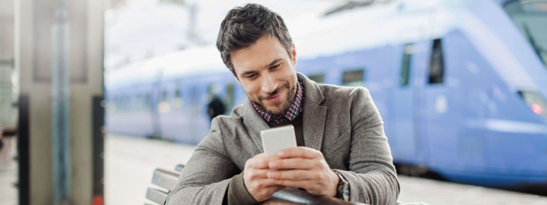 Mann sitzt mit Smartphone in der Hand auf einer Bank am Bahnhof