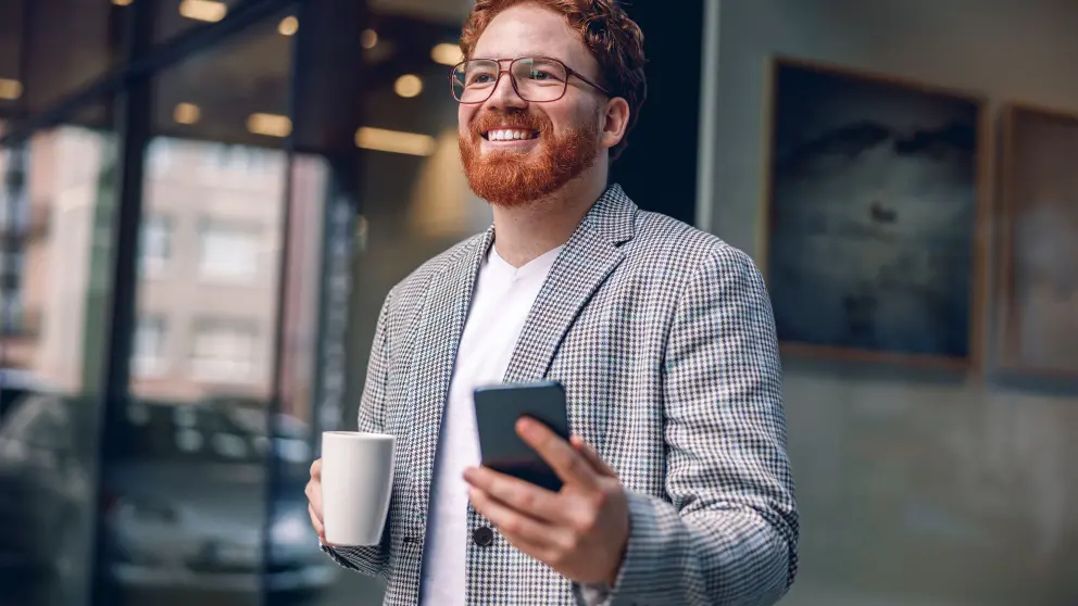 Mann mit roten Haaren, Bart und Brille hält in der einen Hand eine Kaffeetasse und in der anderen Hand ein Smartphone und lächelt