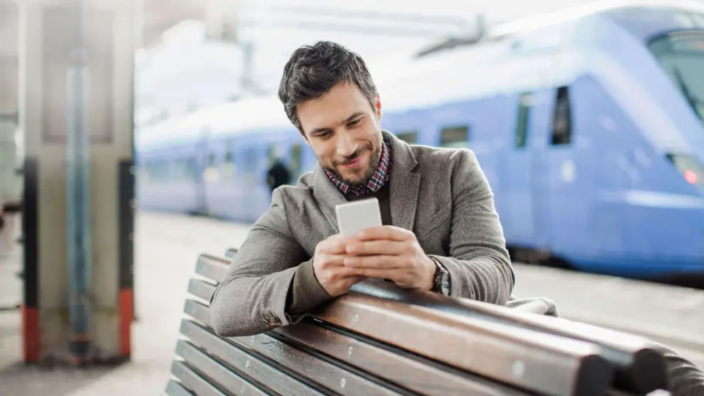 Mann sitzt mit Smartphone in der Hand auf einer Bank am Bahnhof