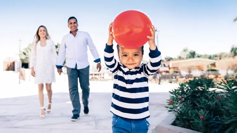 Kind hält roten Ball in den Händen, im Hintergrund laufen seine Eltern Hand in Hand