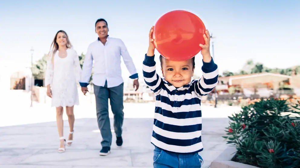Kind hält roten Ball in den Händen, im Hintergrund laufen seine Eltern Hand in Hand