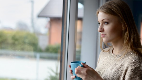 Junge Frau mit Tasse blickt nachdenkich aus dem Fenster