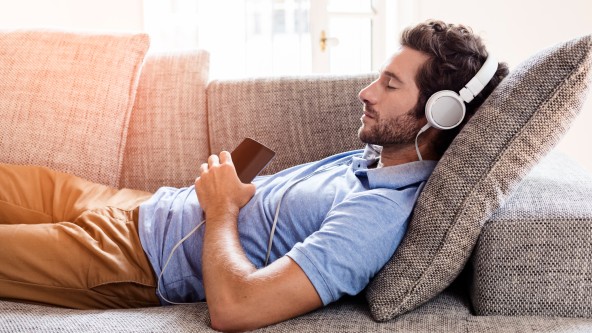 Junger Mann mit Bart liegt mit Kopfhörern auf dem Sofa