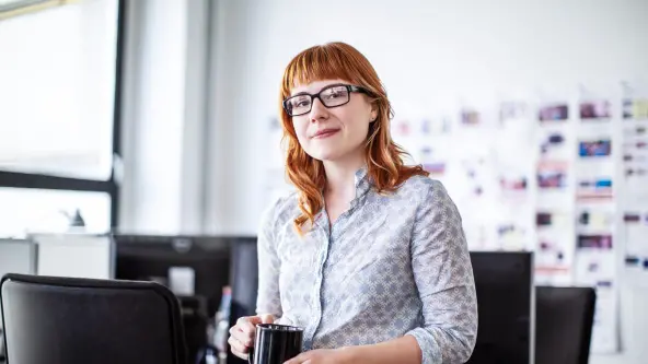 Junge rothaarige Frau mit Brille und einer Tasse in der Hand lächelt