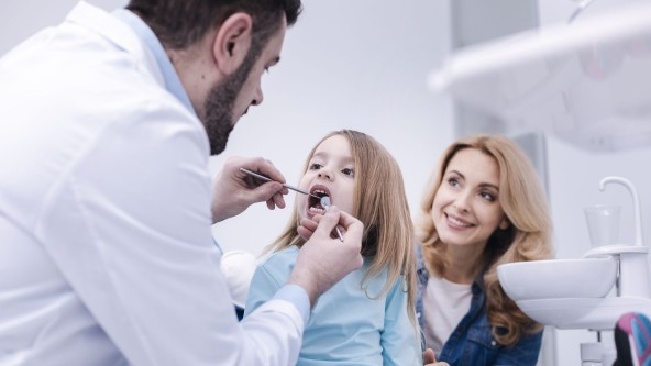 Kind sitzt mit Mutter beim Zahnarzt und wird an den Zähnen untersucht