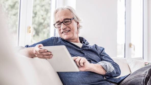 Älterer Mann mit Brille sitzt auf Sofa mit Tablet in der Hand