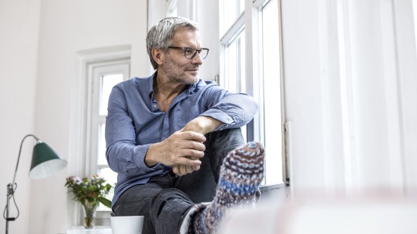 Älterer Mann mit Brille und grauen Haaren sitzt auf einem Fensterbrett